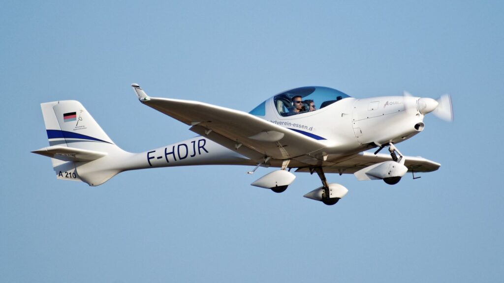 Weiße Aquila A210 mit blauen Streifen und dem Kennzeichen F-HOJR im Flug vor blauem Himmel. Im Cockpit sitzen zwei Personen.
