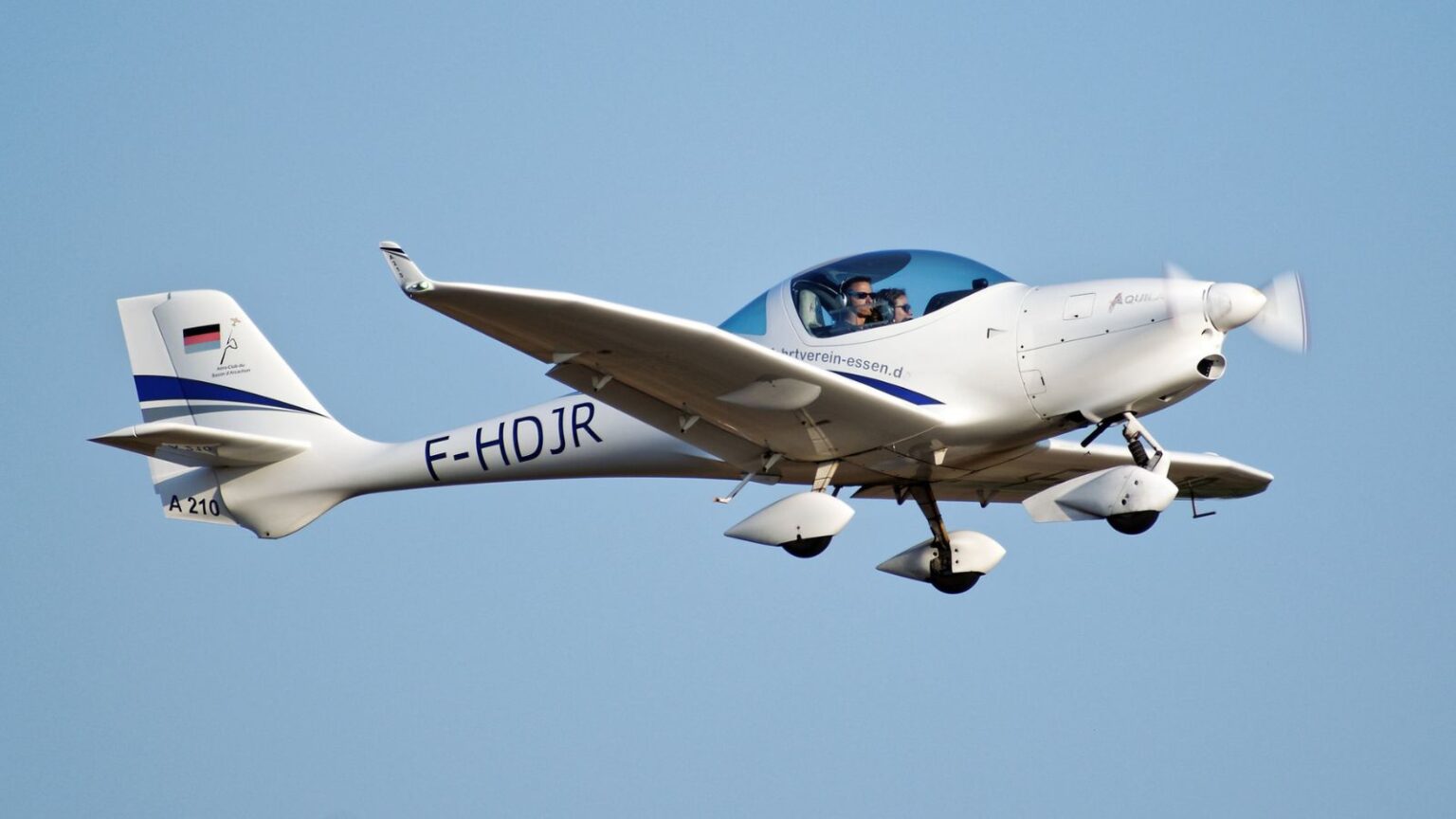 Weiße Aquila A210 mit blauen Streifen und dem Kennzeichen F-HOJR im Flug vor blauem Himmel. Im Cockpit sitzen zwei Personen.